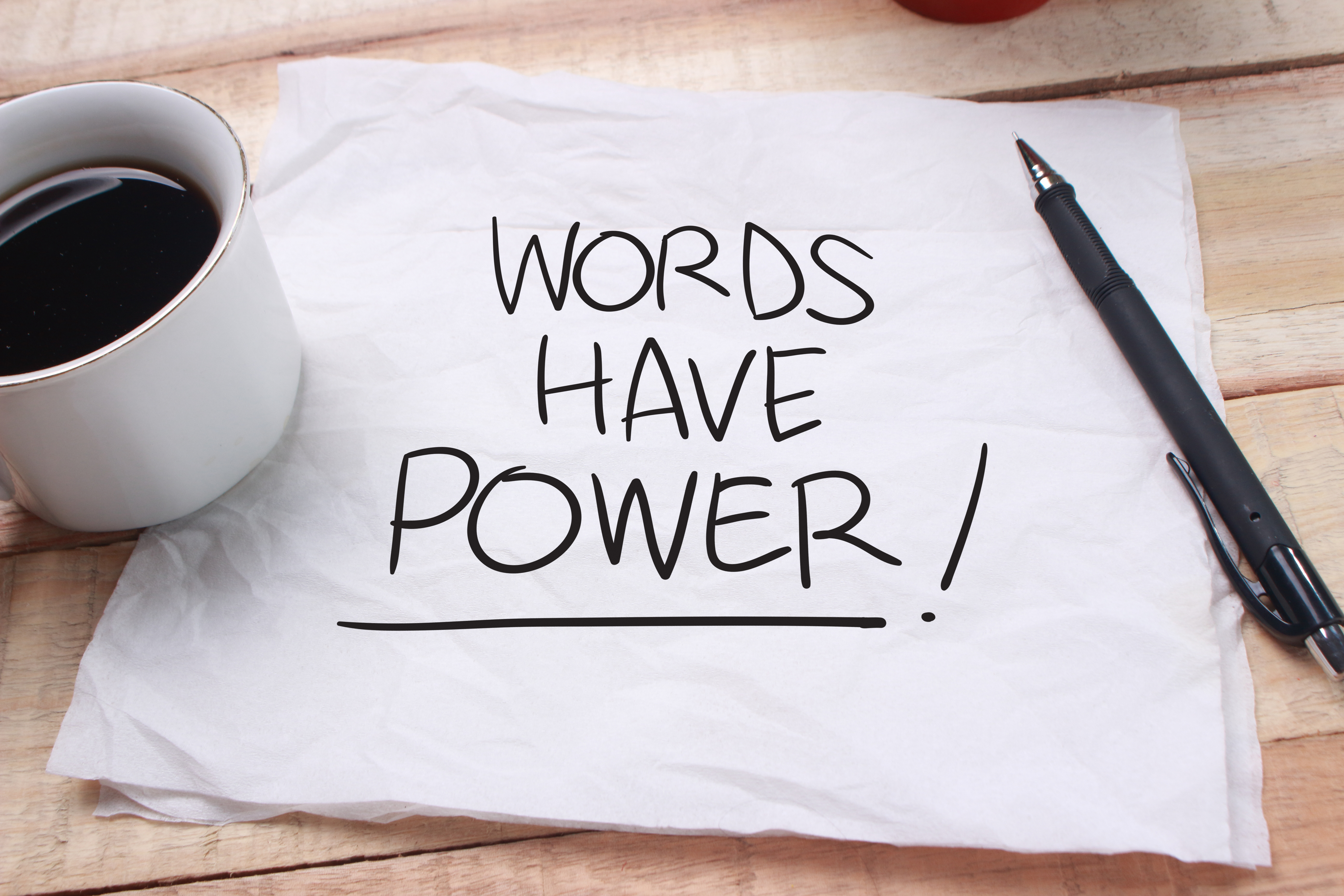A cup of coffee and a pen are placed on a paper napkin with Words have Power! written on it.